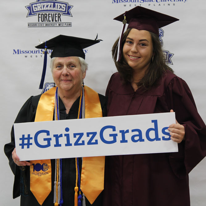 Two MSU-West Plains graduates take a photo at the selfie station holding a sign that reads hashtag Grizz Grads