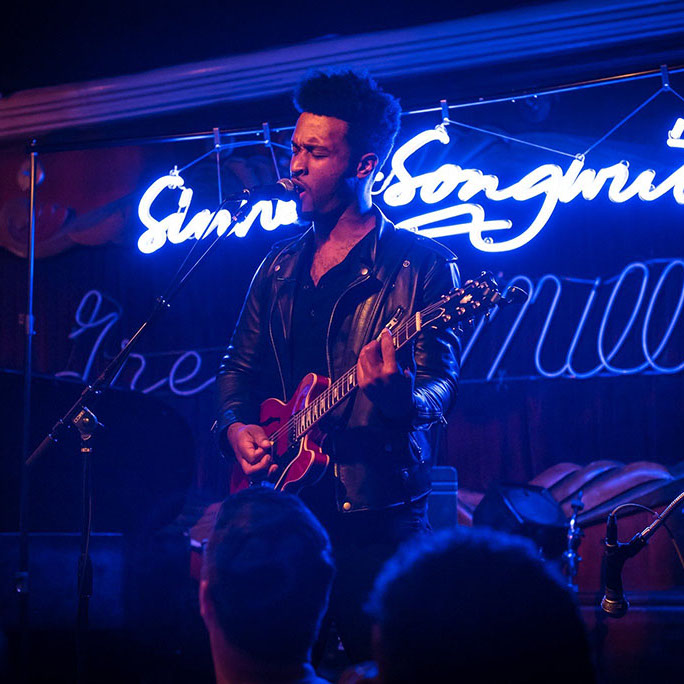 Phillip-Michael Scales performs guitar and singing at a concert with a neon light in the background