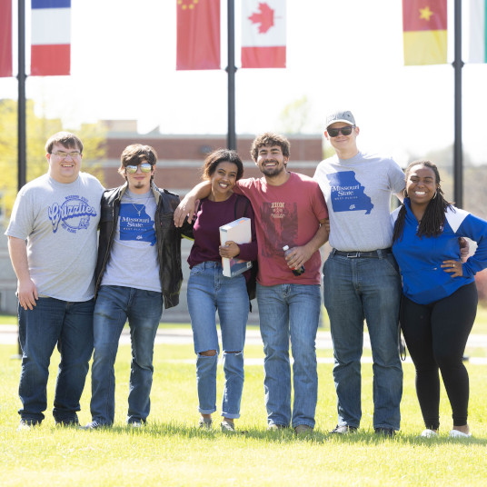 Six MSU-West Plains students wearing Grizzly Gear pose for a picture in the diversity commons with various countries' flags in the background