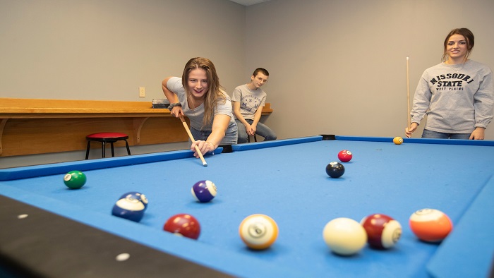 Two women are playing pool while another woman watches from a chair. The woman playing is watching as her appoint takes a shot into a group of pool balls. 