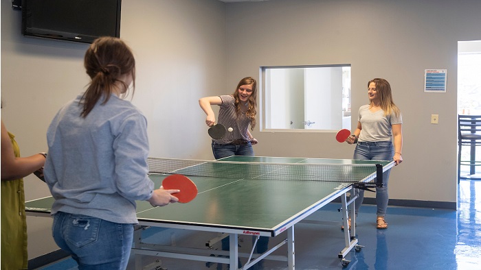 Four women are smiling while playing ping-pong. Three of the women are waiting as the fourth woman hits the ping-pong ball with her paddle. 