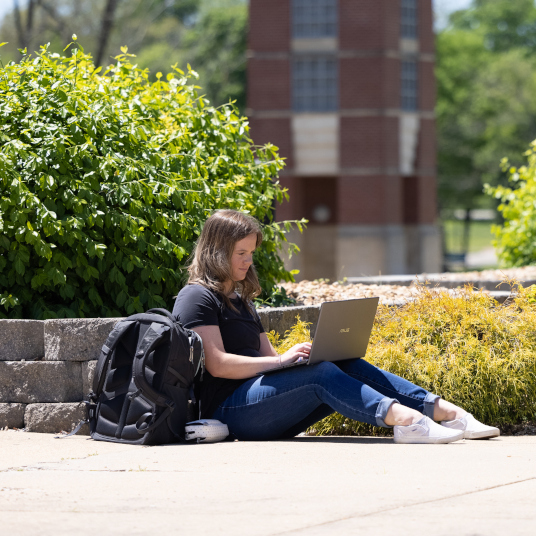 Student sitting on campus with a laptop with the bell tower in the background