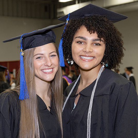 Two graduates smile wearing the caps and gowns